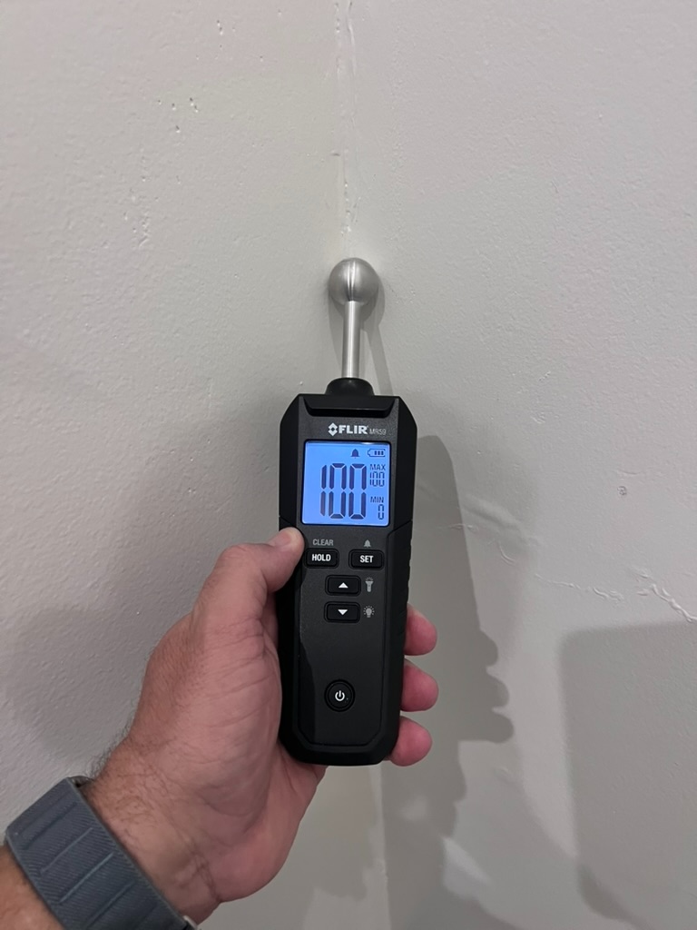 Pin-type moisture meter reading one hundred percent on an interior wall